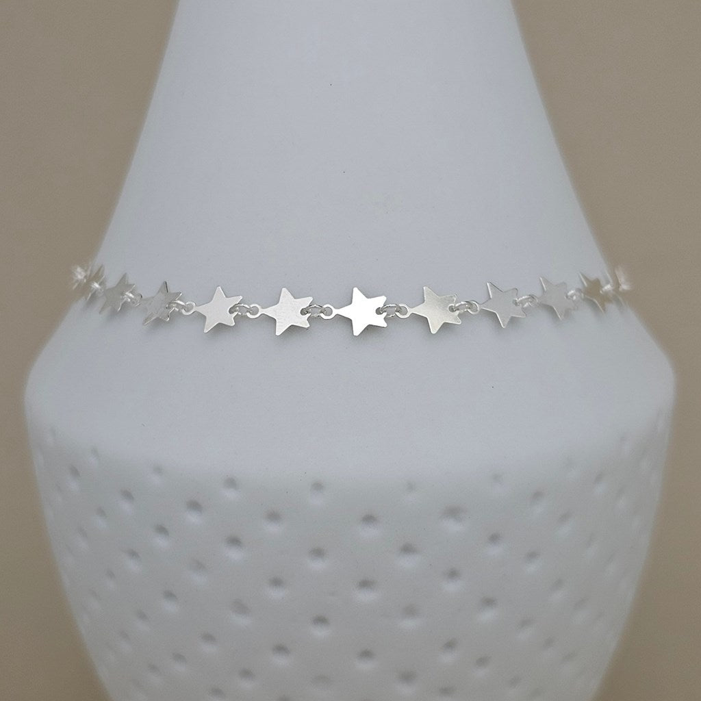 A delicate silver star charm anklet displayed on a white textured cone jewelry stand against a neutral beige background.