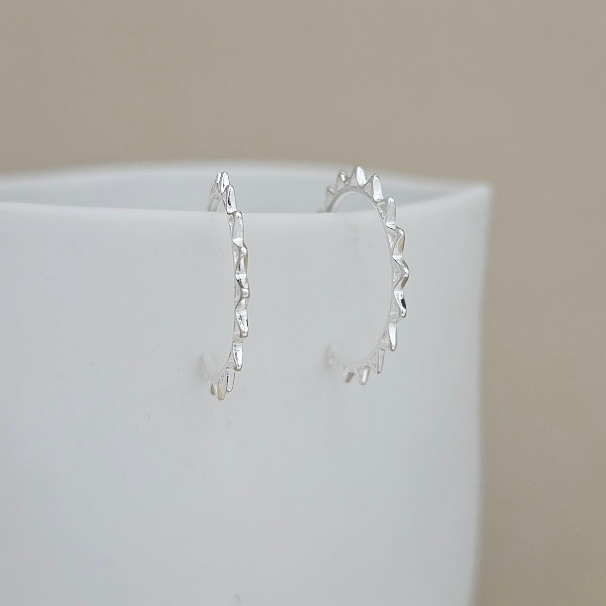 A pair of delicate silver hoop earrings with a textured, scalloped edge, displayed on a white ceramic surface against a soft beige background.