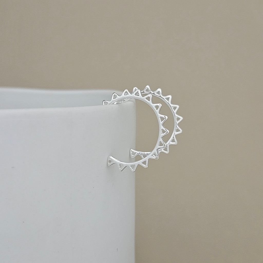 A silver crescent earring with triangular sunburst details on the ends, shown resting on the edge of a white cylindrical display block against a neutral beige background.