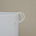 A silver crescent earring with triangular sunburst details on the ends, shown resting on the edge of a white cylindrical display block against a neutral beige background.