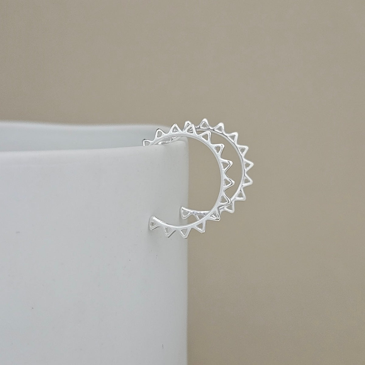 A silver crescent earring with triangular sunburst details on the ends, shown resting on the edge of a white cylindrical display block against a neutral beige background.