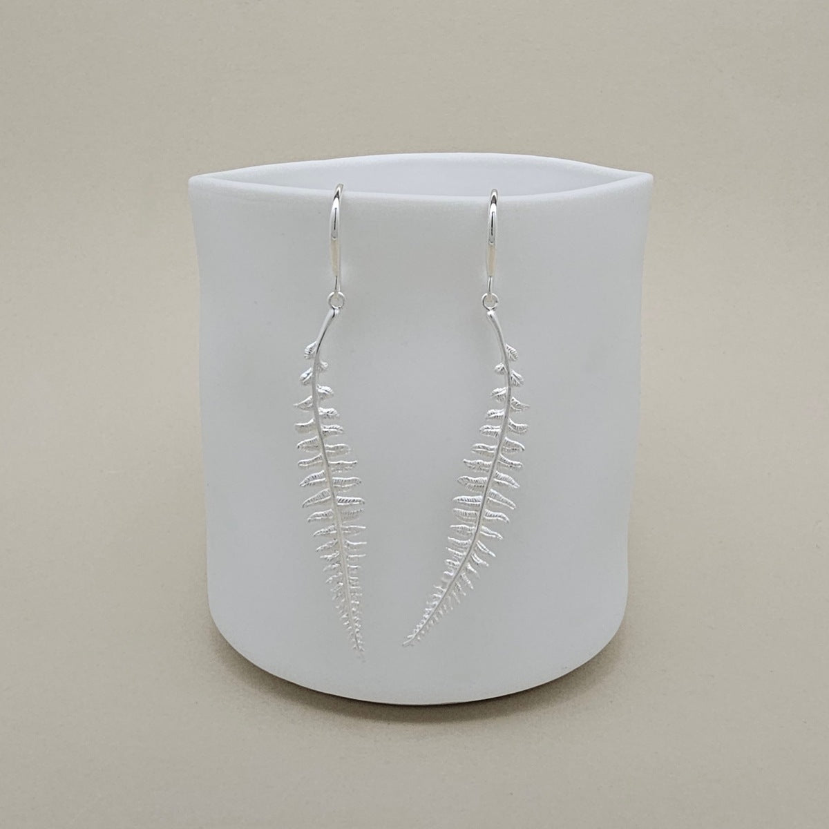 Silver leaf-shaped earrings on a white cylindrical container against a beige background