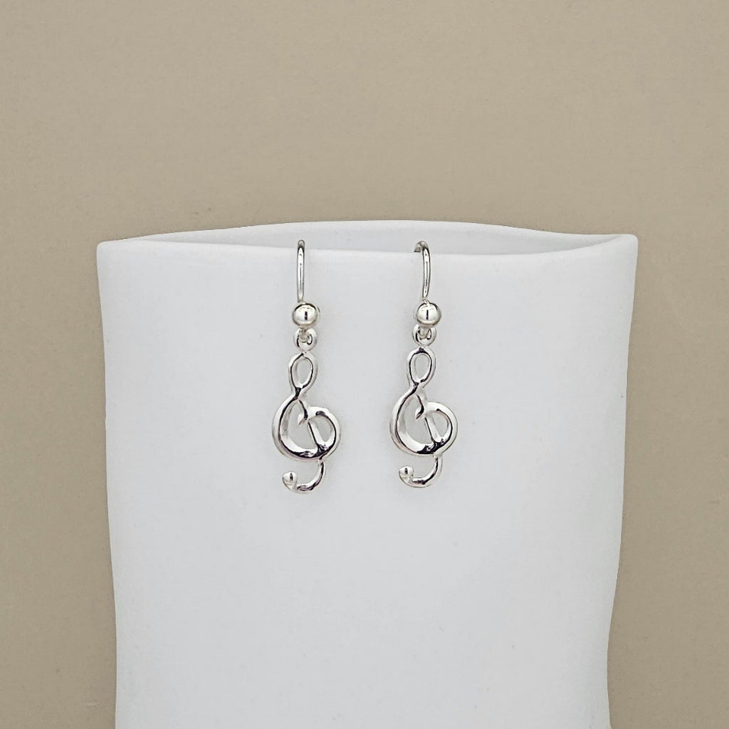 A pair of silver drop earrings featuring interlocking treble clefs, displayed on a white jewelry bust against a solid beige background.