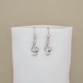 A pair of silver drop earrings featuring interlocking treble clefs, displayed on a white jewelry bust against a solid beige background.