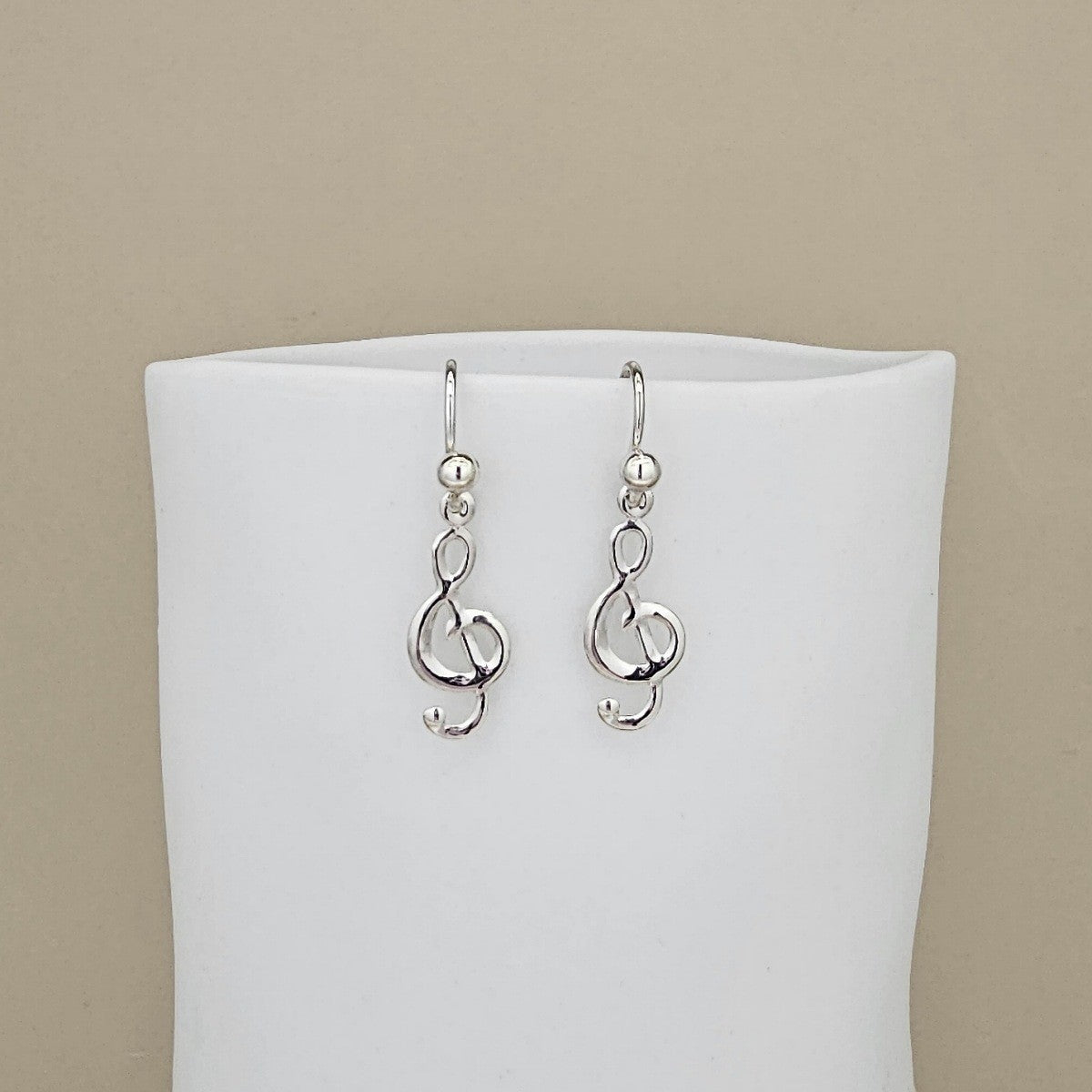 A pair of silver drop earrings featuring interlocking treble clefs, displayed on a white jewelry bust against a solid beige background.