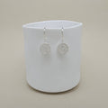 A pair of silver circular drop earrings with intricate filigree detailing, displayed on a white ceramic cup against a warm beige background.