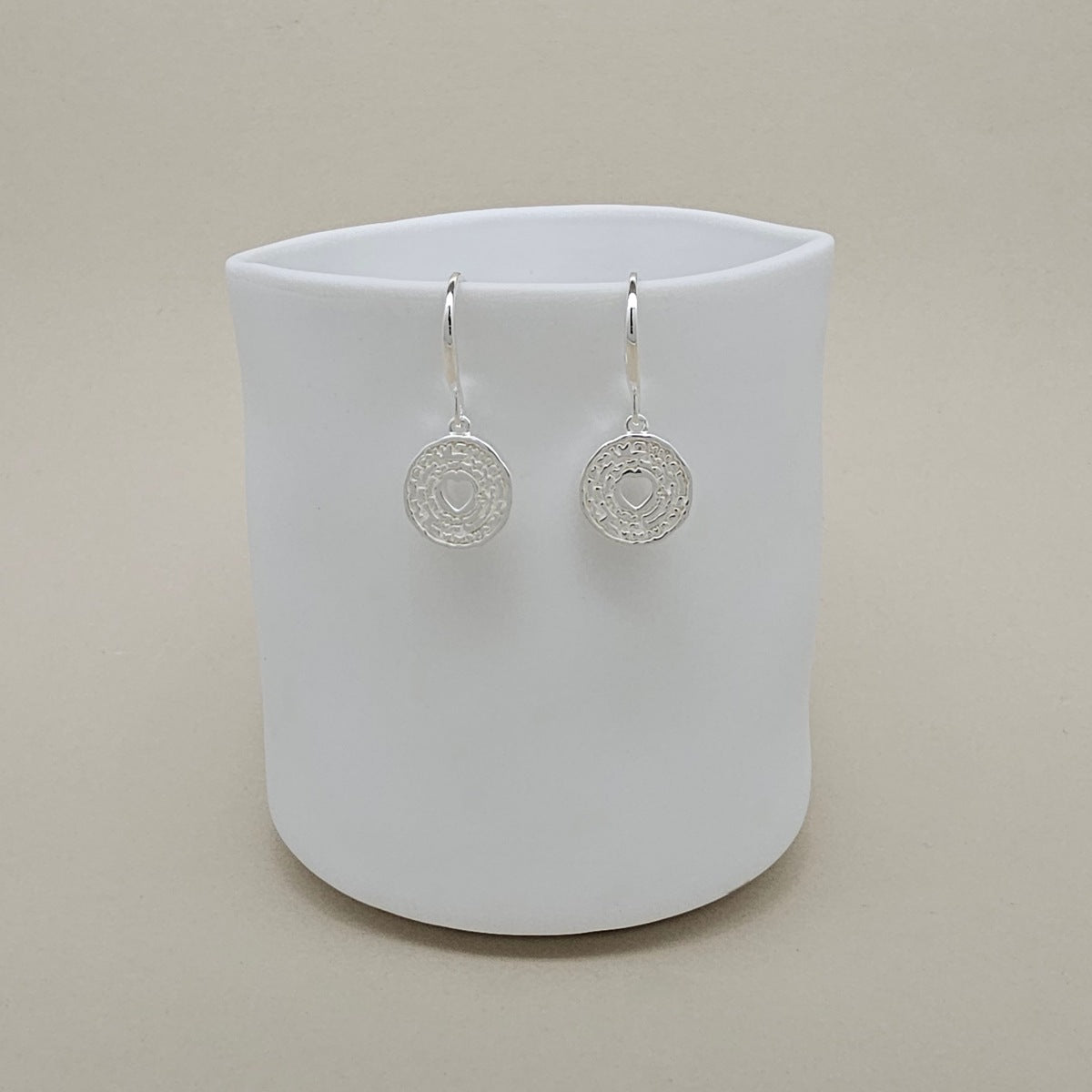 A pair of silver circular drop earrings with intricate filigree detailing, displayed on a white ceramic cup against a warm beige background.