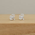 A pair of silver tree stud earrings displayed on a light wooden surface against a neutral beige background.