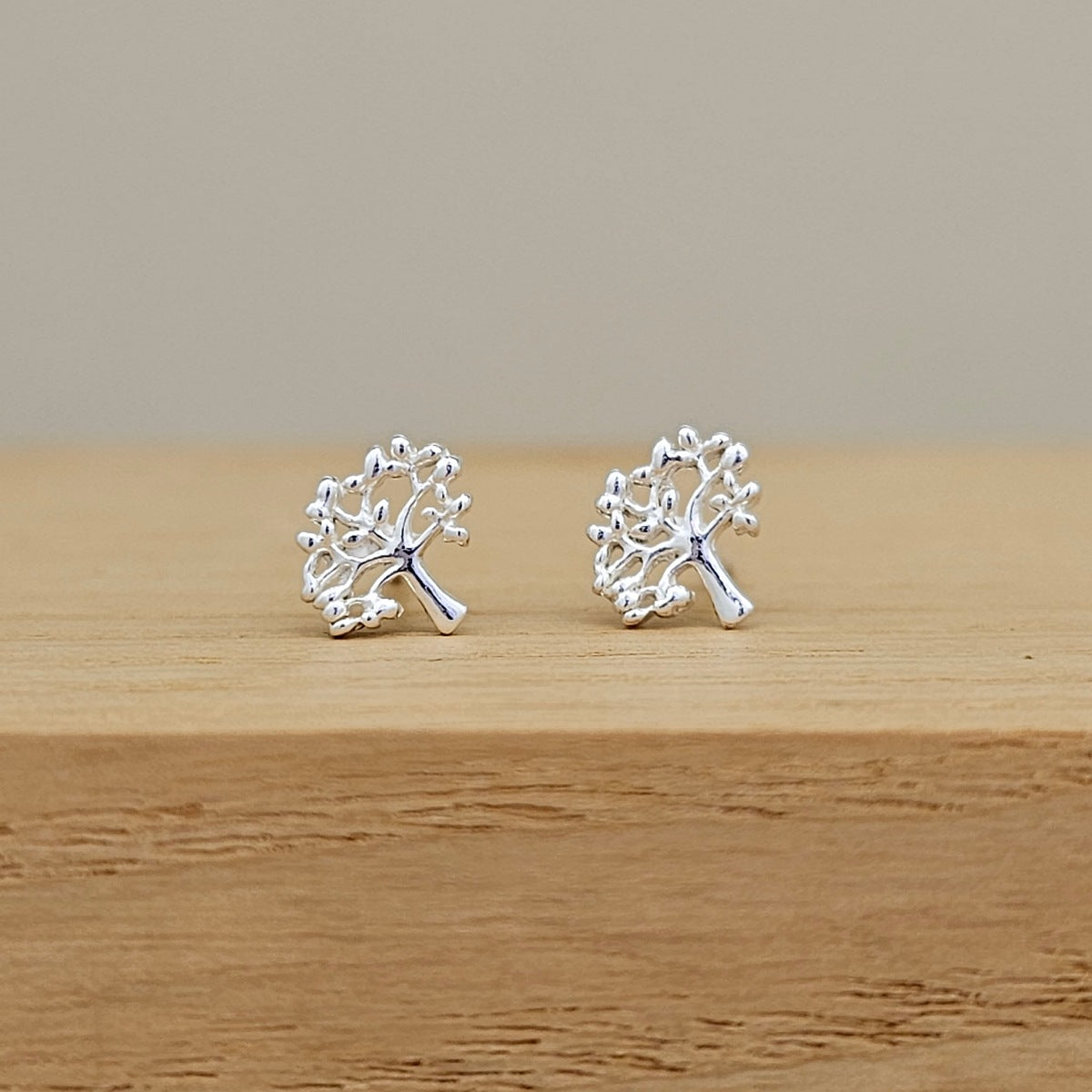 A pair of silver tree stud earrings displayed on a light wooden surface against a neutral beige background.