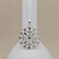 A silver filigree pendant necklace featuring a circular design with cutout hearts, flowers, and leaves, hanging from a delicate silver chain on a white display bust against a warm beige background.