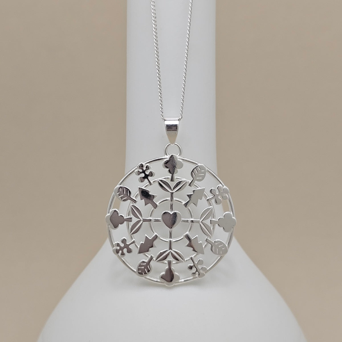 A silver filigree pendant necklace featuring a circular design with cutout hearts, flowers, and leaves, hanging from a delicate silver chain on a white display bust against a warm beige background.