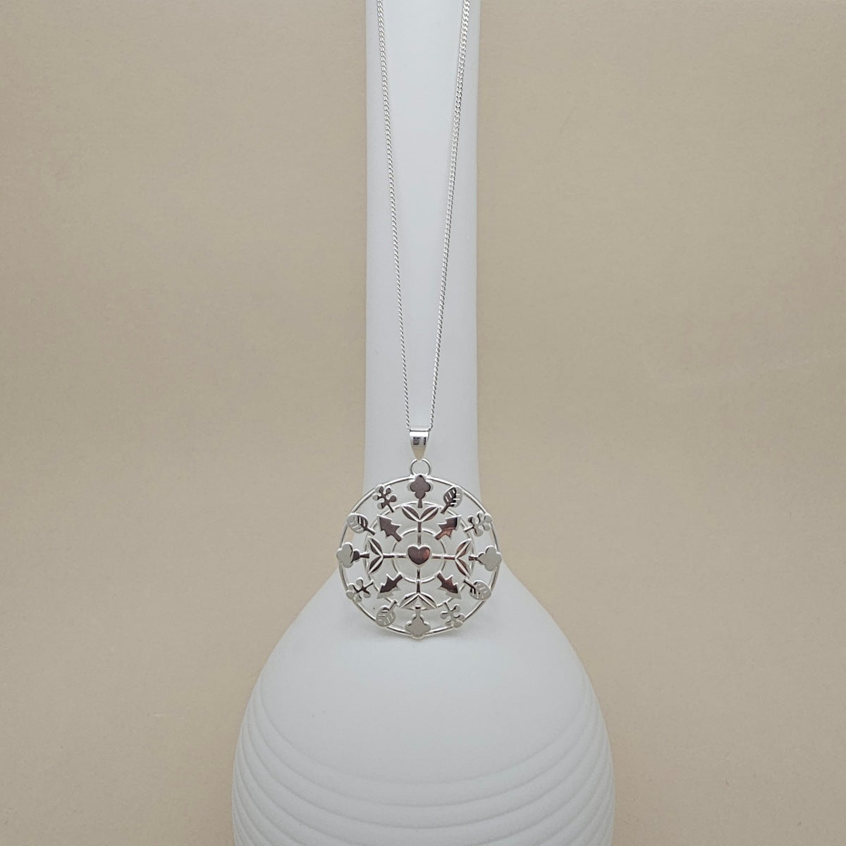 A silver-toned circular pendant necklace with an intricate cutout floral design, hanging from a delicate chain on a white display stand against a warm beige background.