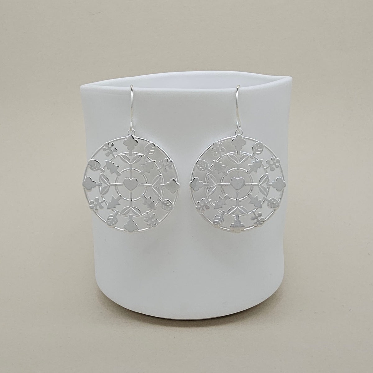 A pair of silver filigree dangle earrings with circular cutout patterns, hanging from silver fishhook wires, displayed against a white ceramic cup on a beige background.