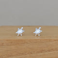 A pair of silver stud earrings shaped like cartoon turtles, resting on a light brown wooden surface against a neutral gray background.