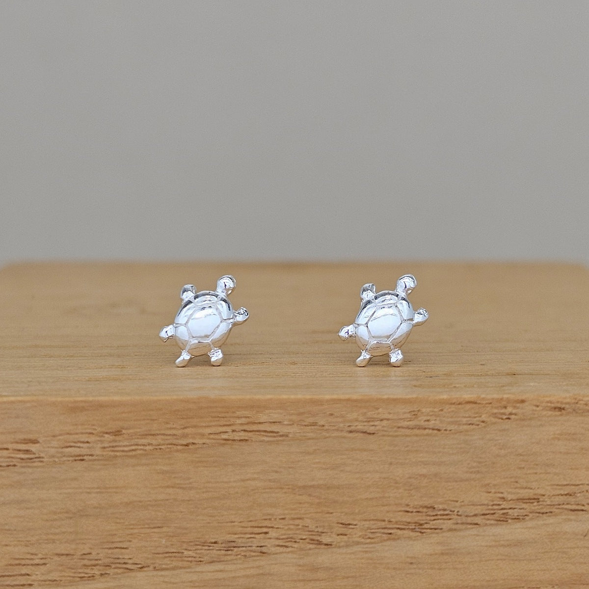 A pair of silver stud earrings shaped like cartoon turtles, resting on a light brown wooden surface against a neutral gray background.