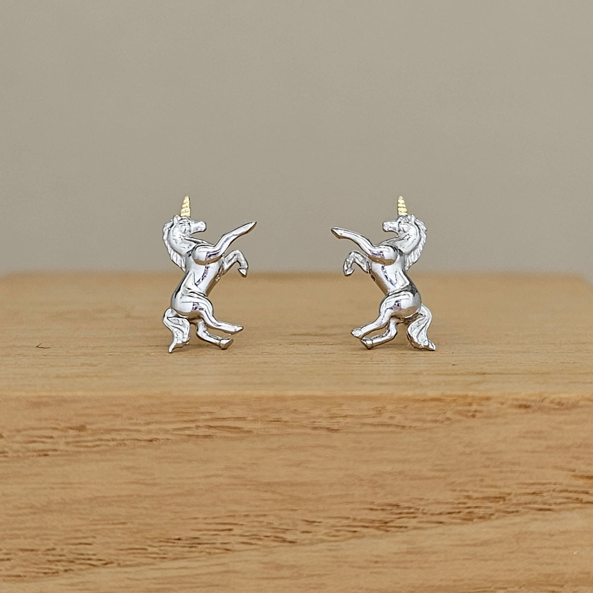 A pair of sterling silver unicorn stud earrings with gold-colored horns, displayed on a wooden block against a neutral beige background.