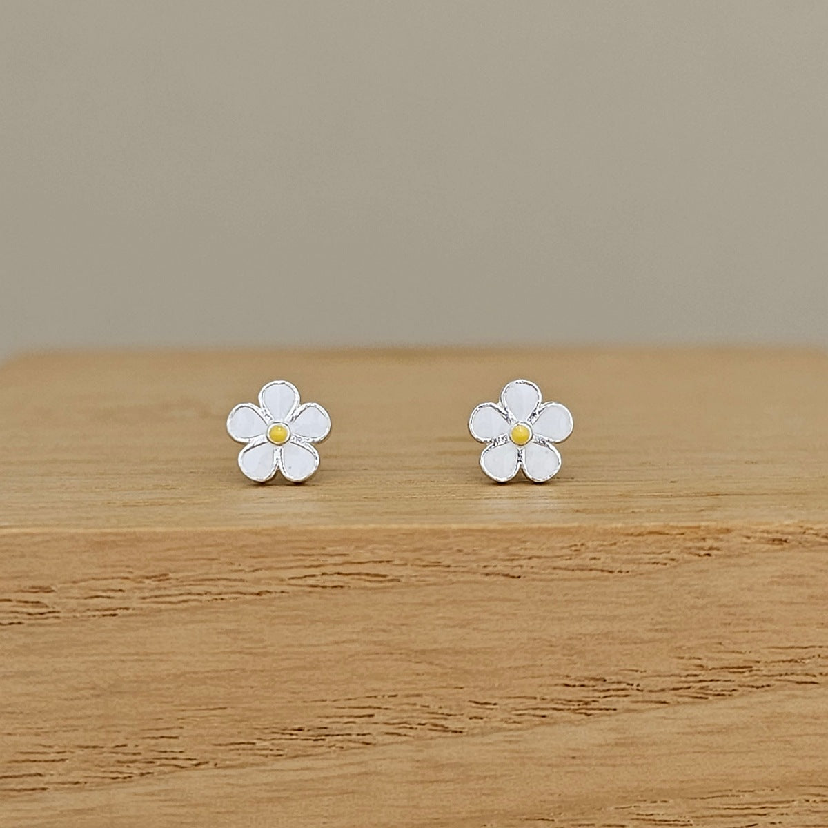 A pair of small white flower stud earrings with yellow centers, displayed on a light wooden surface against a neutral beige background.