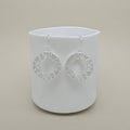 A pair of silver-toned circular drop earrings with a textured, openwork geometric border, displayed on a matte white cylindrical stand against a warm beige background.
