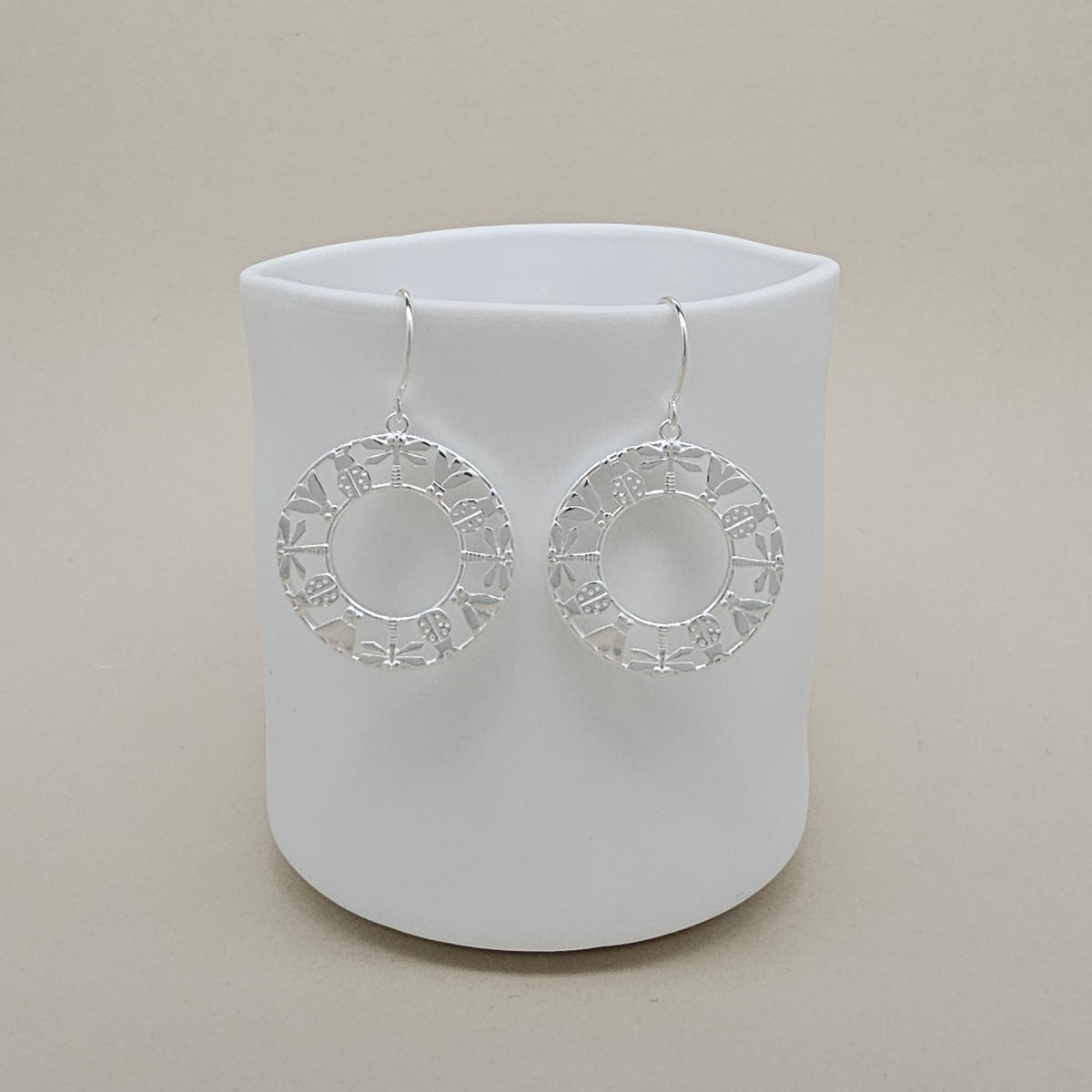 A pair of silver-toned circular drop earrings with a textured, openwork geometric border, displayed on a matte white cylindrical stand against a warm beige background.