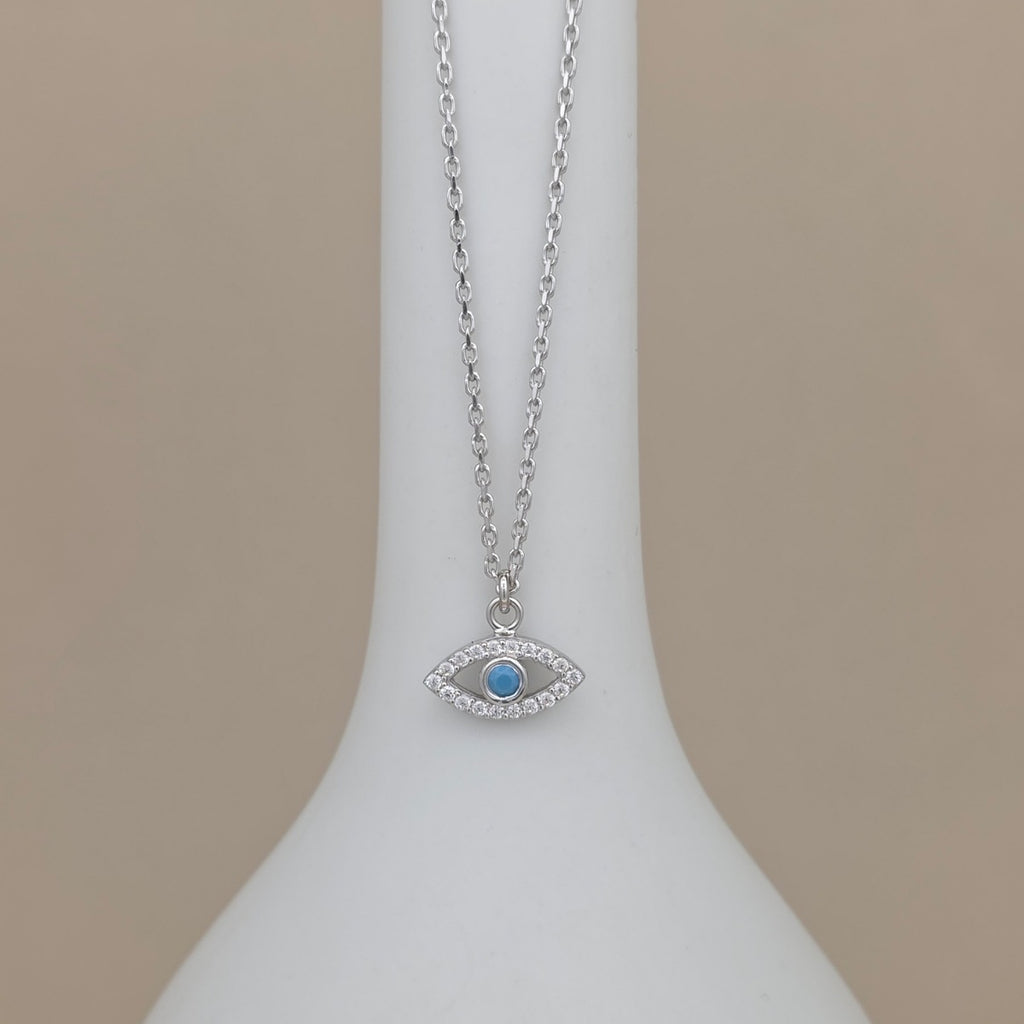 A silver-tone evil eye pendant necklace with a light blue center stone, displayed on a white jewelry bust against a warm beige background.