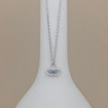 A silver-tone evil eye pendant necklace with a light blue center stone, displayed on a white jewelry bust against a warm beige background.