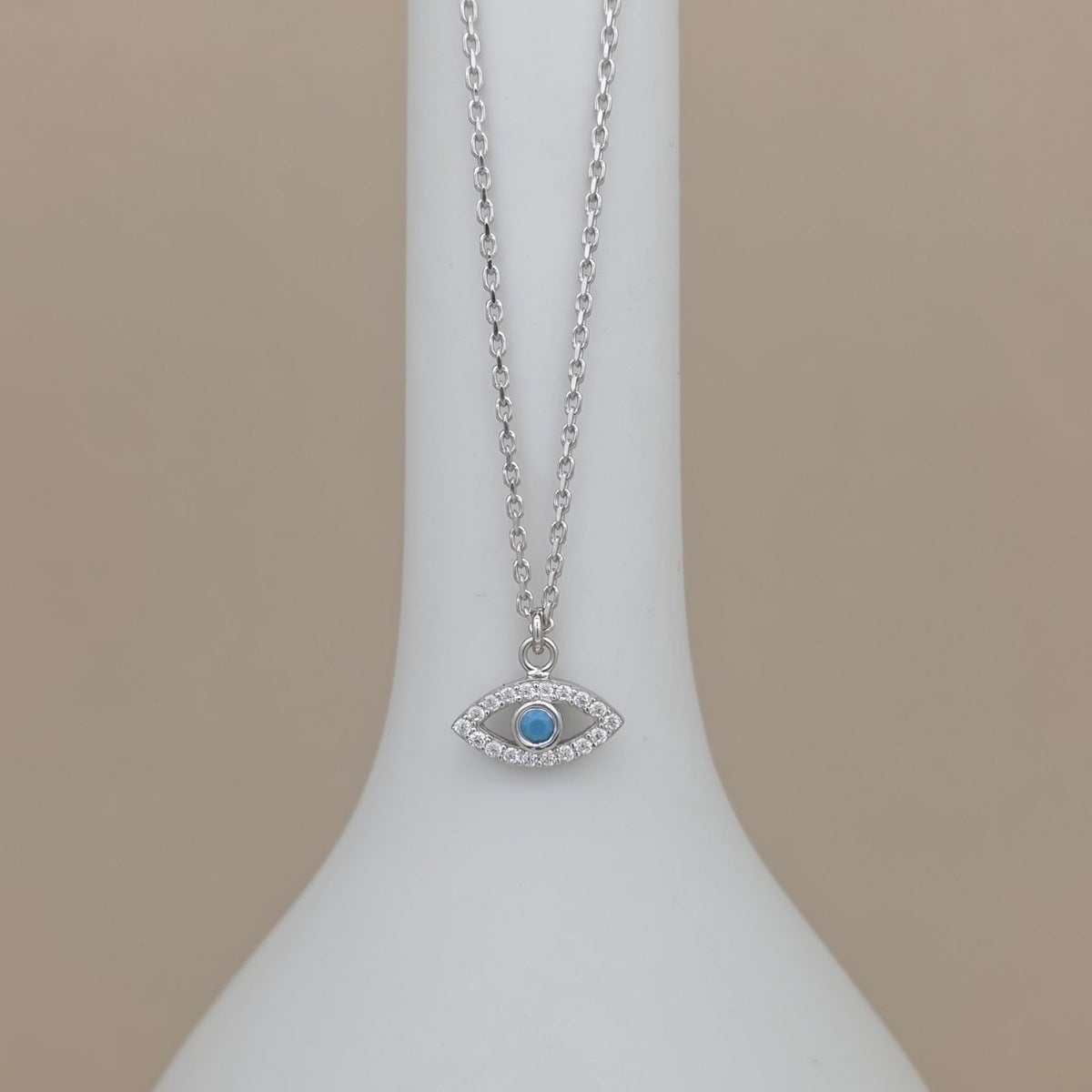 A silver-tone evil eye pendant necklace with a light blue center stone, displayed on a white jewelry bust against a warm beige background.