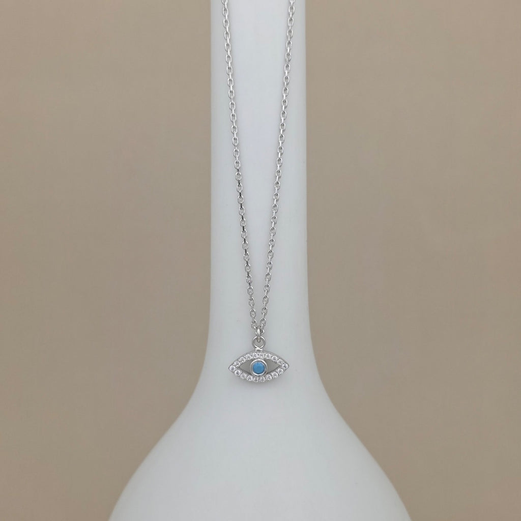 A silver-toned evil eye necklace with a light blue center stone, displayed on a white vase against a beige background.