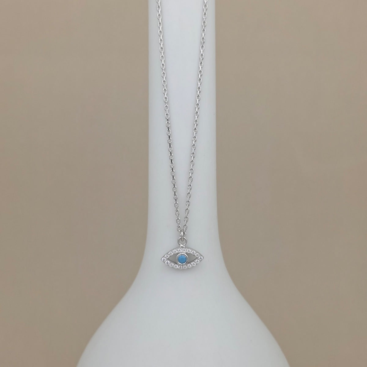 A silver-toned evil eye necklace with a light blue center stone, displayed on a white vase against a beige background.