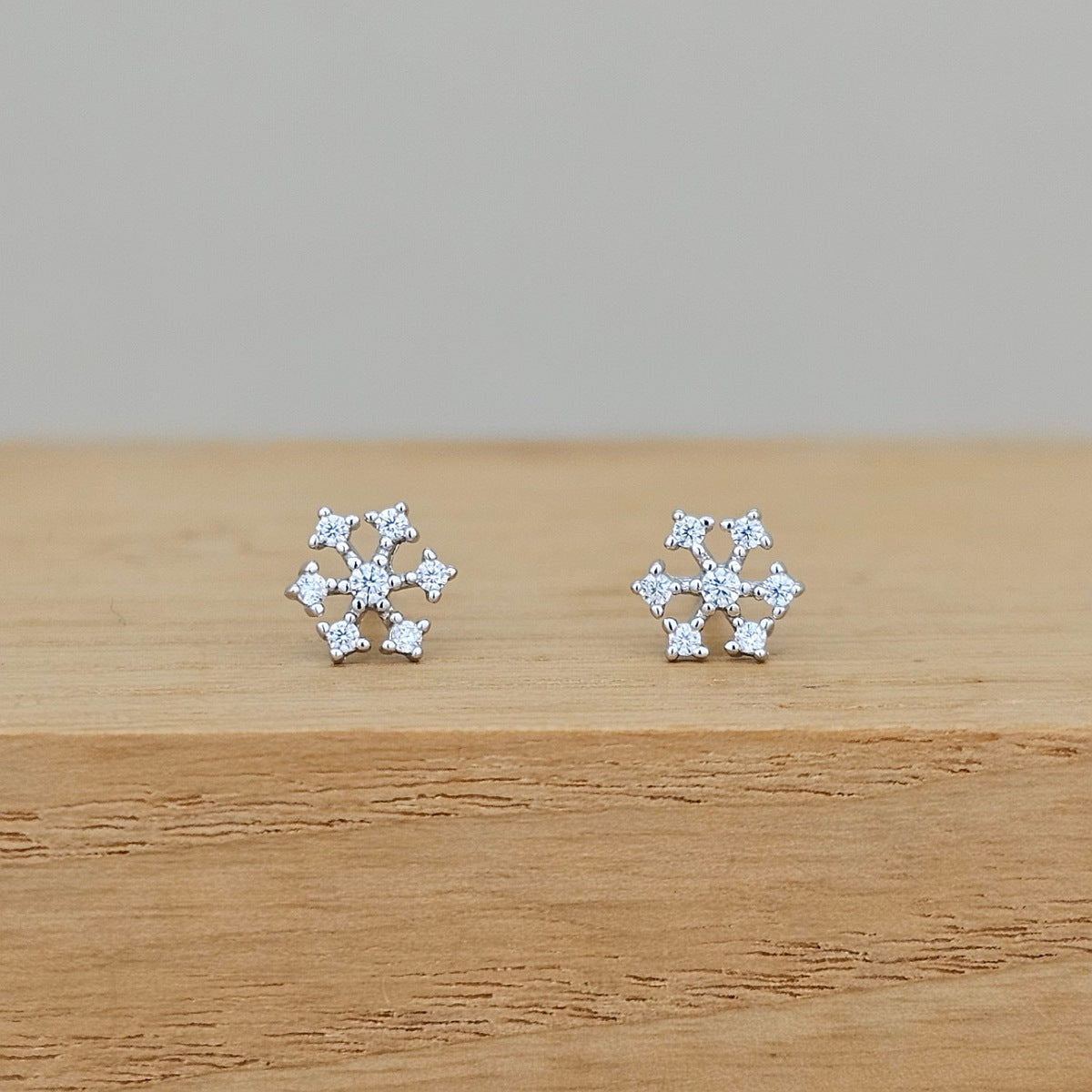 A pair of silver snowflake stud earrings set with small clear stones, resting on a light brown wooden surface against a neutral gray background.