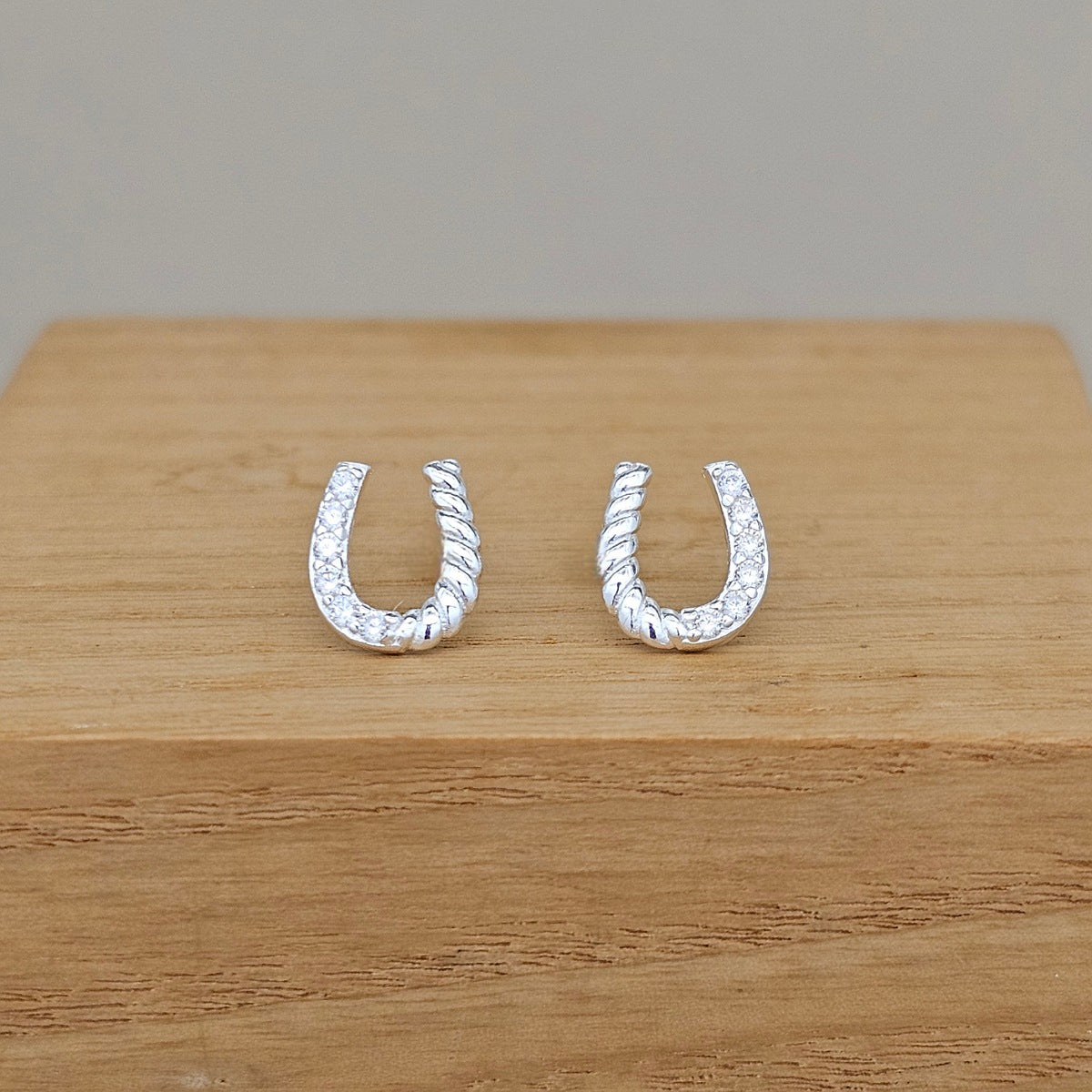 A pair of silver U-shaped hoop earrings with a twisted rope texture and small clear stones, displayed on a light wood block against a neutral gray background.