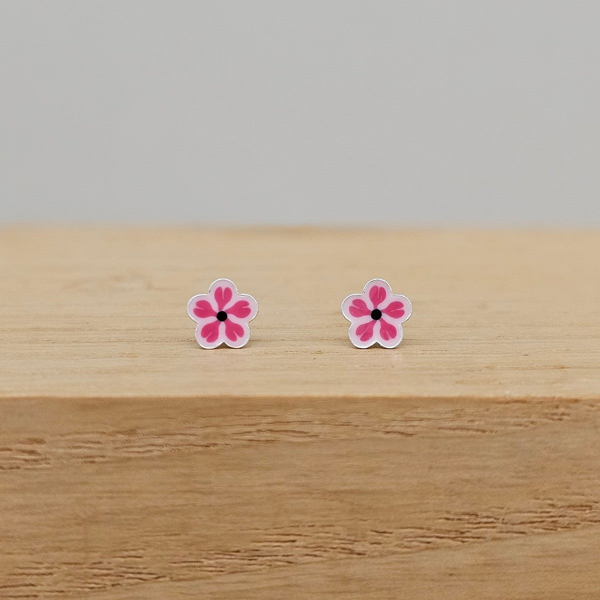 A pair of small flower-shaped stud earrings with pink petals, white outlines, and black centers, resting on a light wood surface against a neutral gray background.