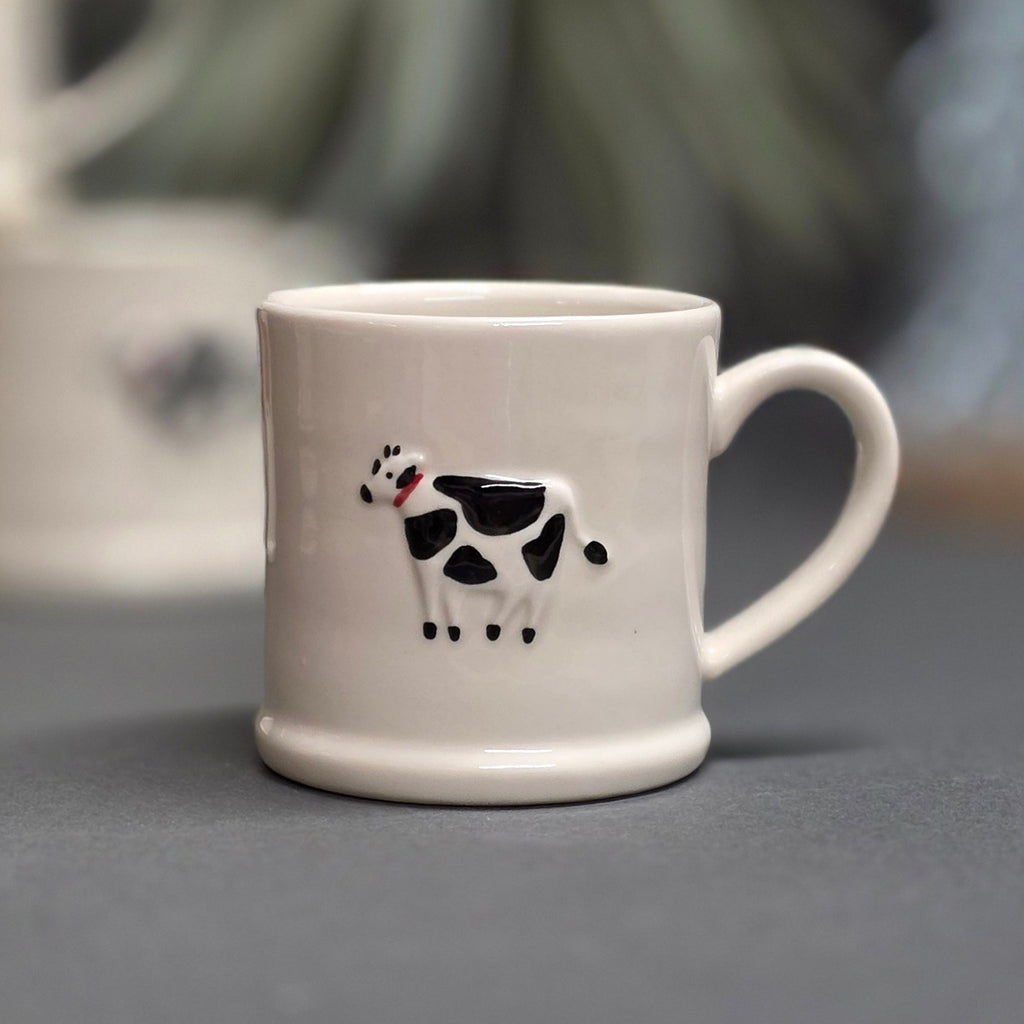 A white ceramic mug with a black and white cow illustration on the front, set against a blurred grey background.