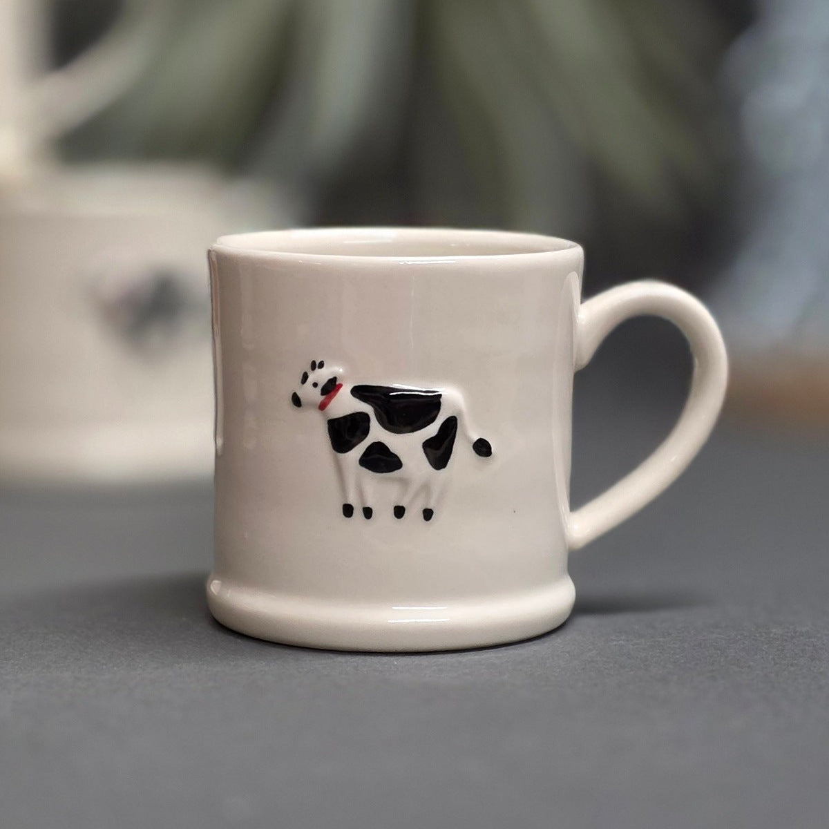 A white ceramic mug with a black and white cow illustration on the front, set against a blurred grey background.