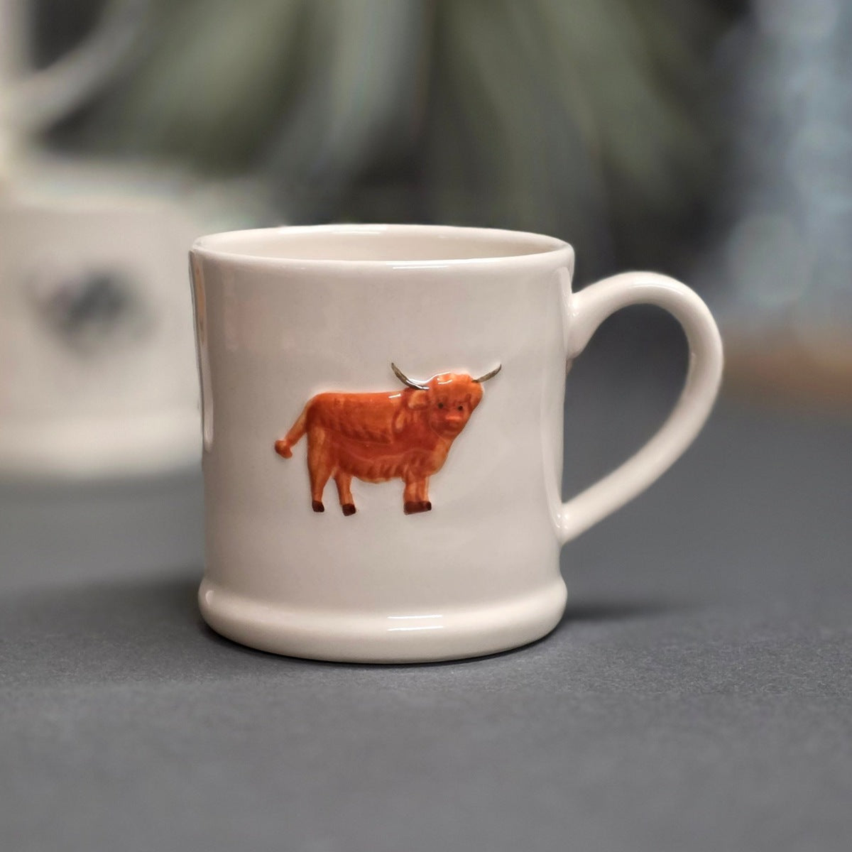 A white ceramic mug featuring a raised, three-dimensional orange Highland cow design on the side, set against a blurred grey background.