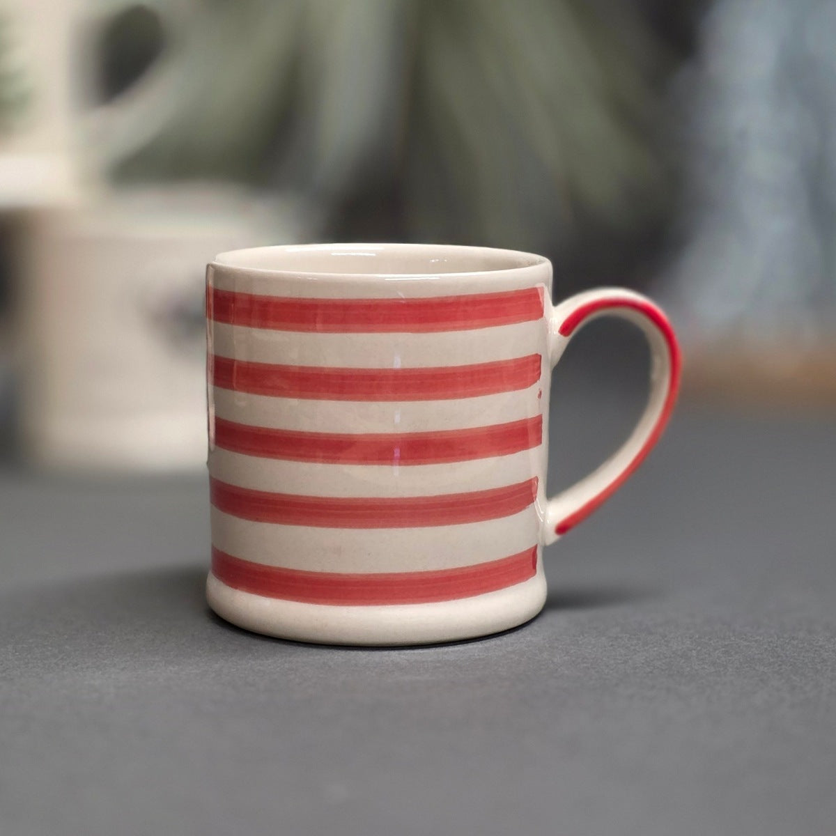 A ceramic mug with horizontal red and white stripes and a red handle, sitting on a dark grey surface with a blurred background.