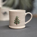 An off-white ceramic mug with a small green pine tree and red berries design on the front, set against a blurred grey background.