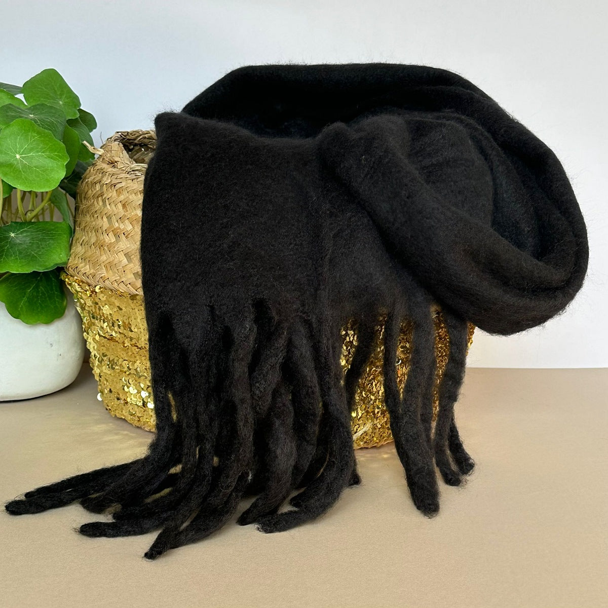 Black chunky soft scarf with tassels draped over a woven basket on a neutral background.
