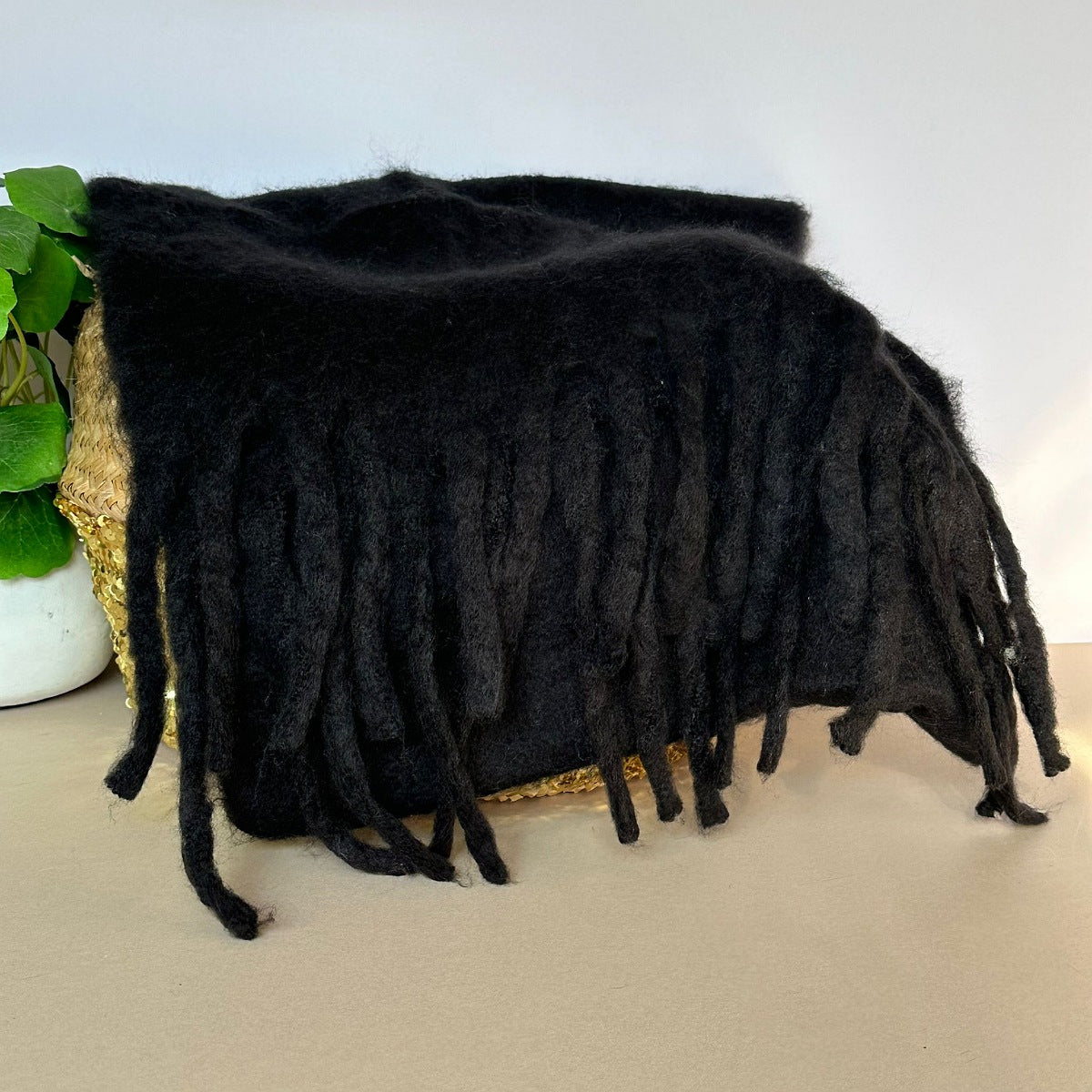 Black fuzzy soft scarf with long fringe draped over a woven basket with a gold sequin front, placed on a tabletop next to a green plant.

