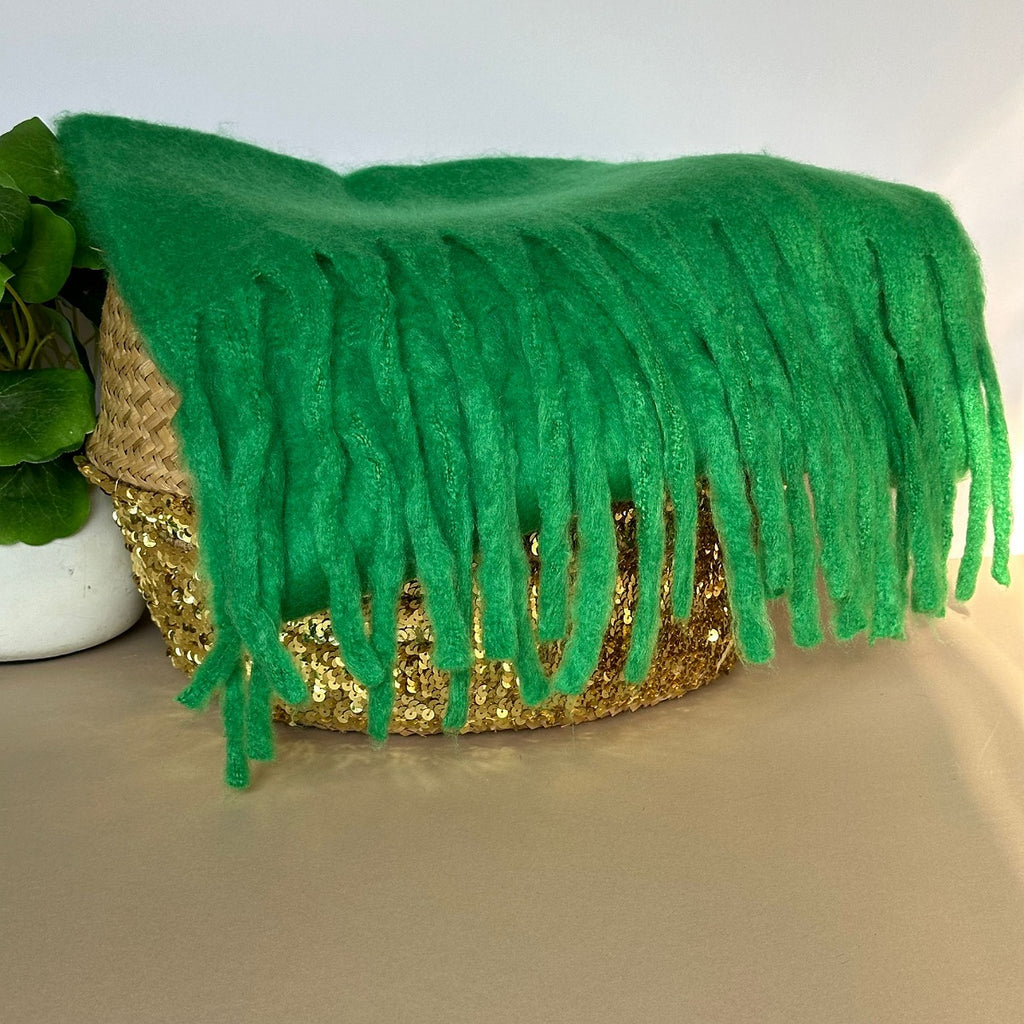 Bright green fuzzy soft scarf with long fringe draped over a woven basket with a gold sequin front, placed on a tabletop next to a green plant.
