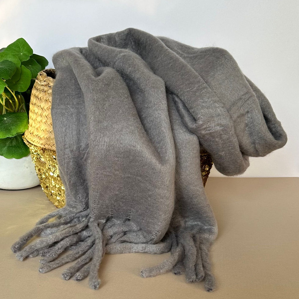 Grey chunky soft scarf with tassels draped over a woven basket on a neutral background.
