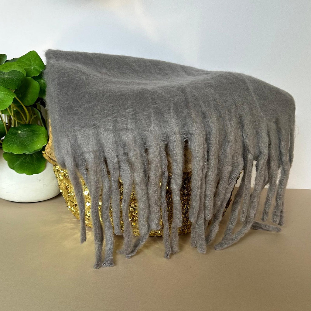 Grey fuzzy soft scarf with long fringe draped over a woven basket with a gold sequin front, placed on a tabletop next to a green plant.
