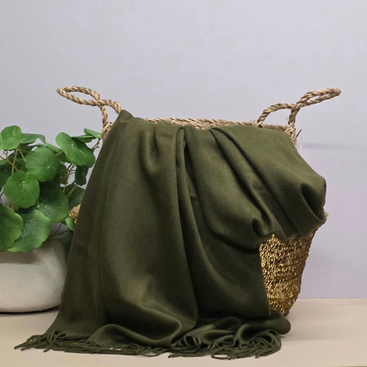 A green fabric with fringed edges draped over a woven straw basket, with a potted green plant on the left against a light grey background.