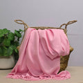 A pink fabric with fringed edges draped over a woven straw basket, with a potted green plant on the left against a light grey background.