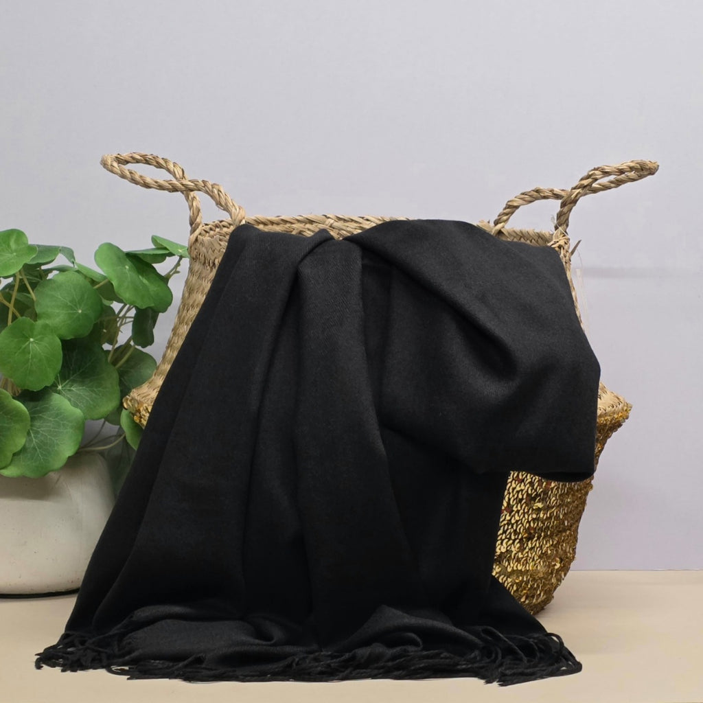 A black woven throw blanket draped over a natural straw basket with rope handles, set on a light beige surface against a pale grey wall, with a potted green plant at the side.