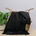 A black woven throw blanket draped over a natural straw basket with rope handles, set on a light beige surface against a pale grey wall, with a potted green plant at the side.