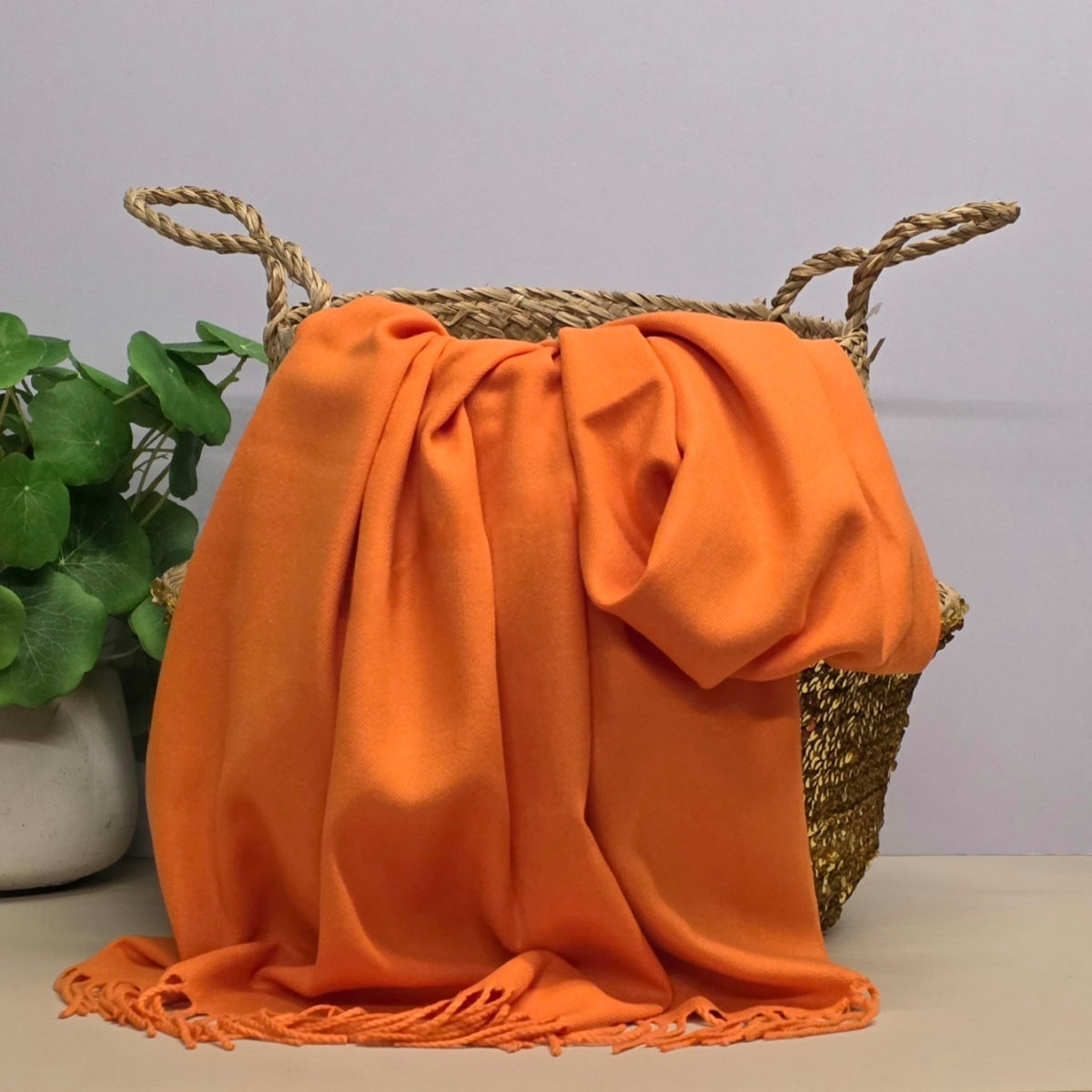 A bright orange fabric with fringed edges draped over a woven straw basket, with a potted green plant on the left against a light grey background.