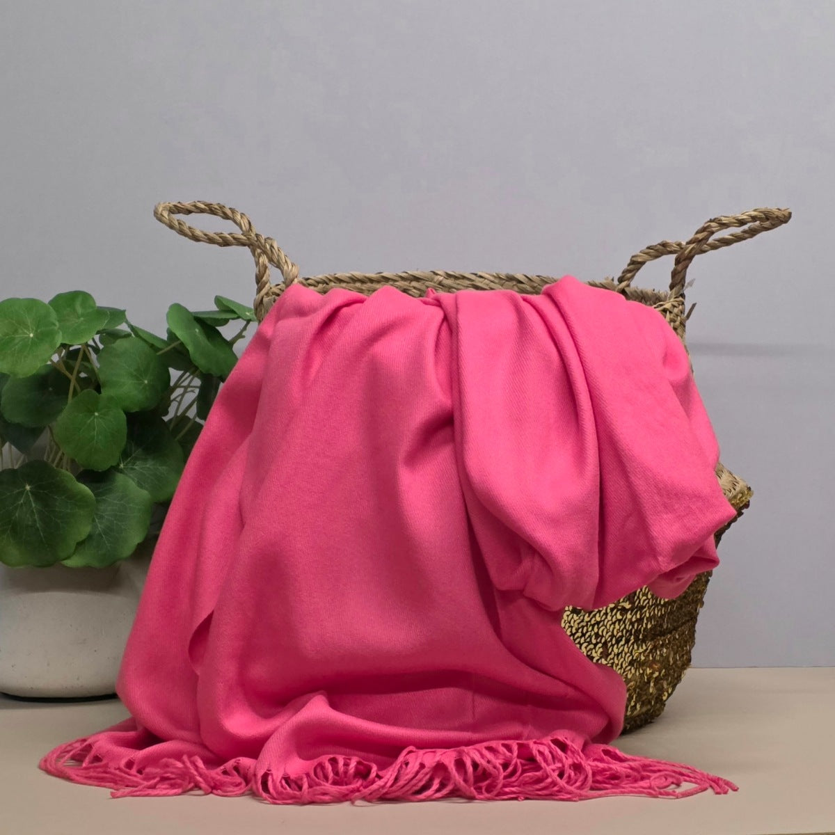A pink fabric with fringed edges draped over a woven straw basket, with a potted green plant on the left against a light grey background.
