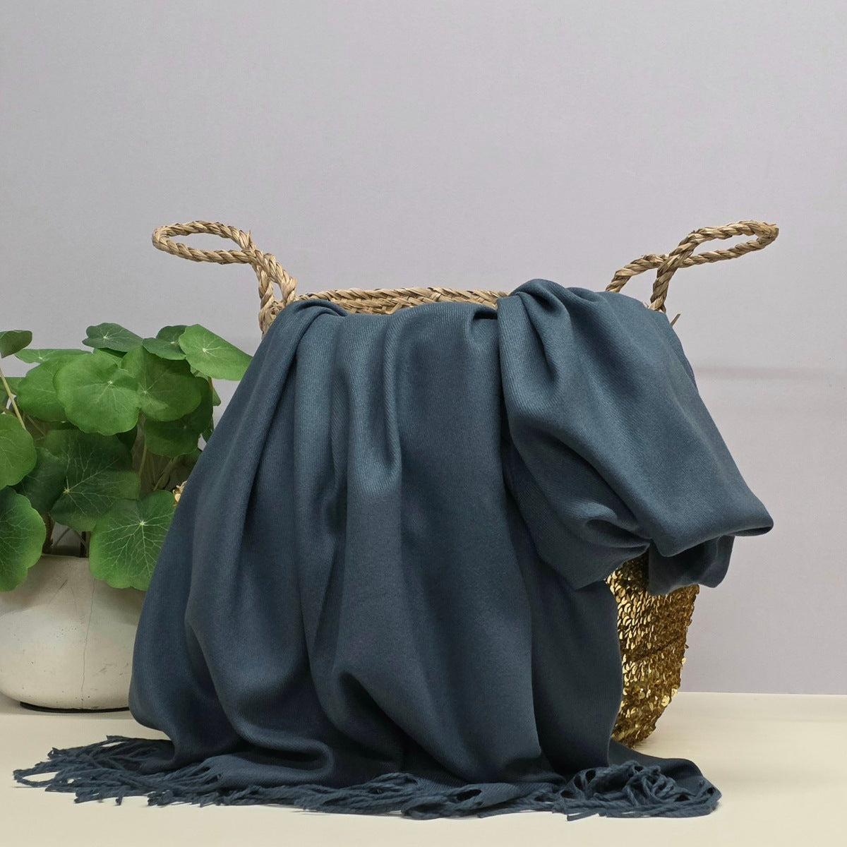 A dark teal blue fringed throw blanket draped over a woven straw basket, with a potted green plant in the background against a light gray wall.
