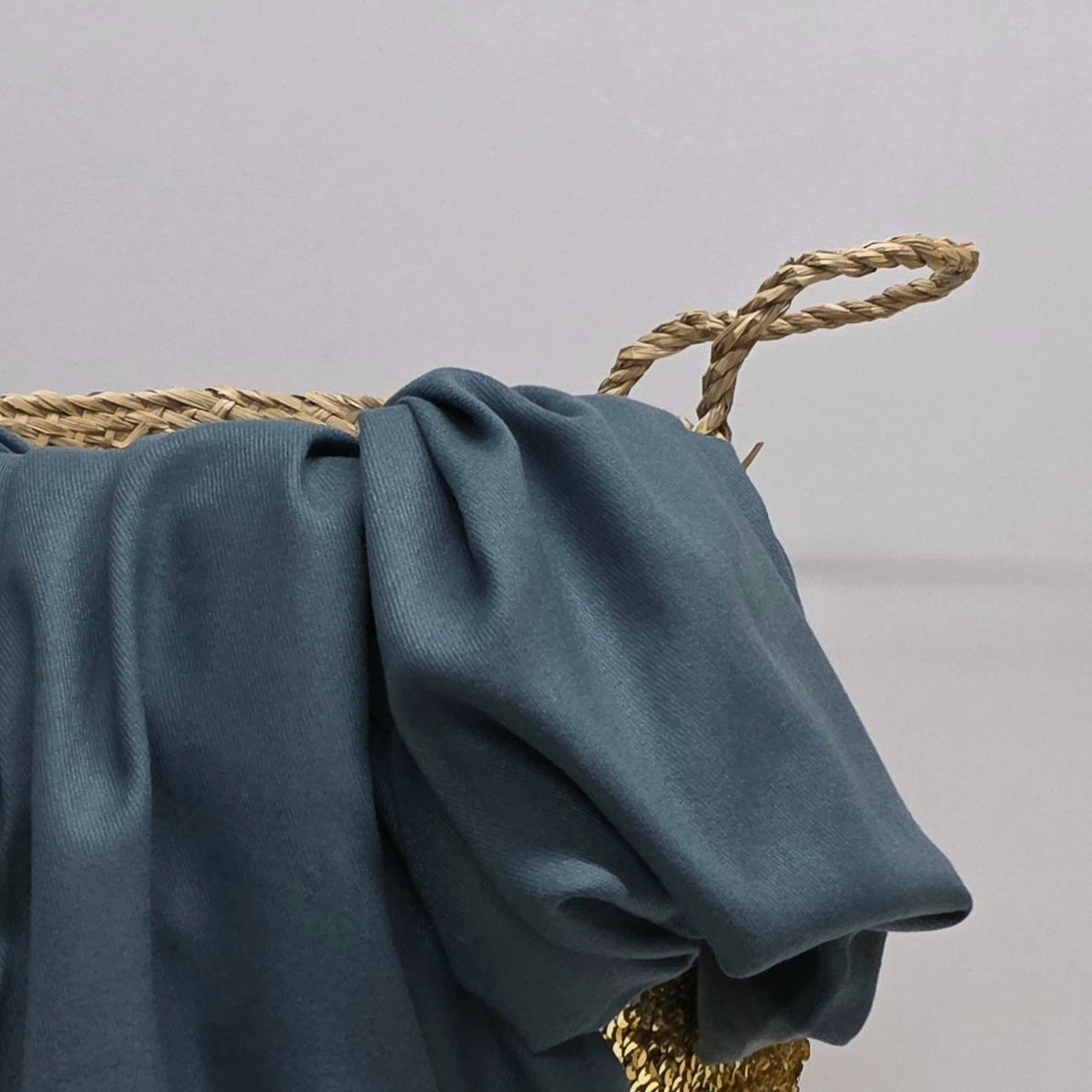 A close-up of a dusty slate blue fabric draped over a woven straw bag handle, showing the soft texture and color of the material.