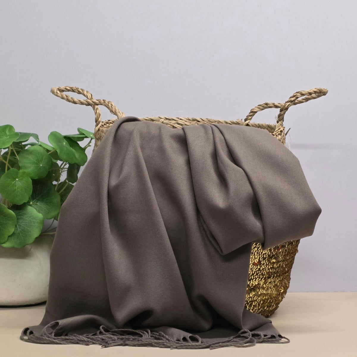 A draped taupe-brown scarf with fringed edges, styled over a woven straw basket with a potted green plant in the background.